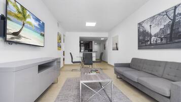 A modern living room with a grey sofa, a coffee table, and a TV on the wall. There is a dining area with a table and chairs in the background.