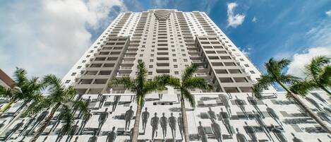 A tall building with a mural of people on its facade, palm trees in front, and a clear blue sky.