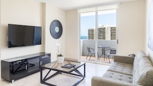 A modern living room with a flat-screen TV, a glass coffee table, a sofa, a potted orchid, and a sliding glass door leading to a balcony with a table and chairs. - Fortune House Hotel Suites (Miami)