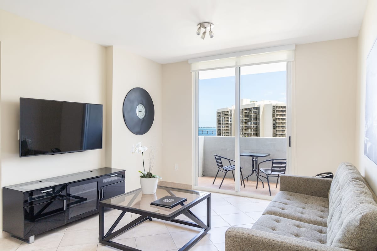 A modern living room with a flat-screen TV, a glass coffee table, a sofa, a potted orchid, and a sliding glass door leading to a balcony with a table and chairs.