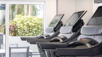 A row of stationary bikes with screens, a window with a view of greenery, and a table with chairs.