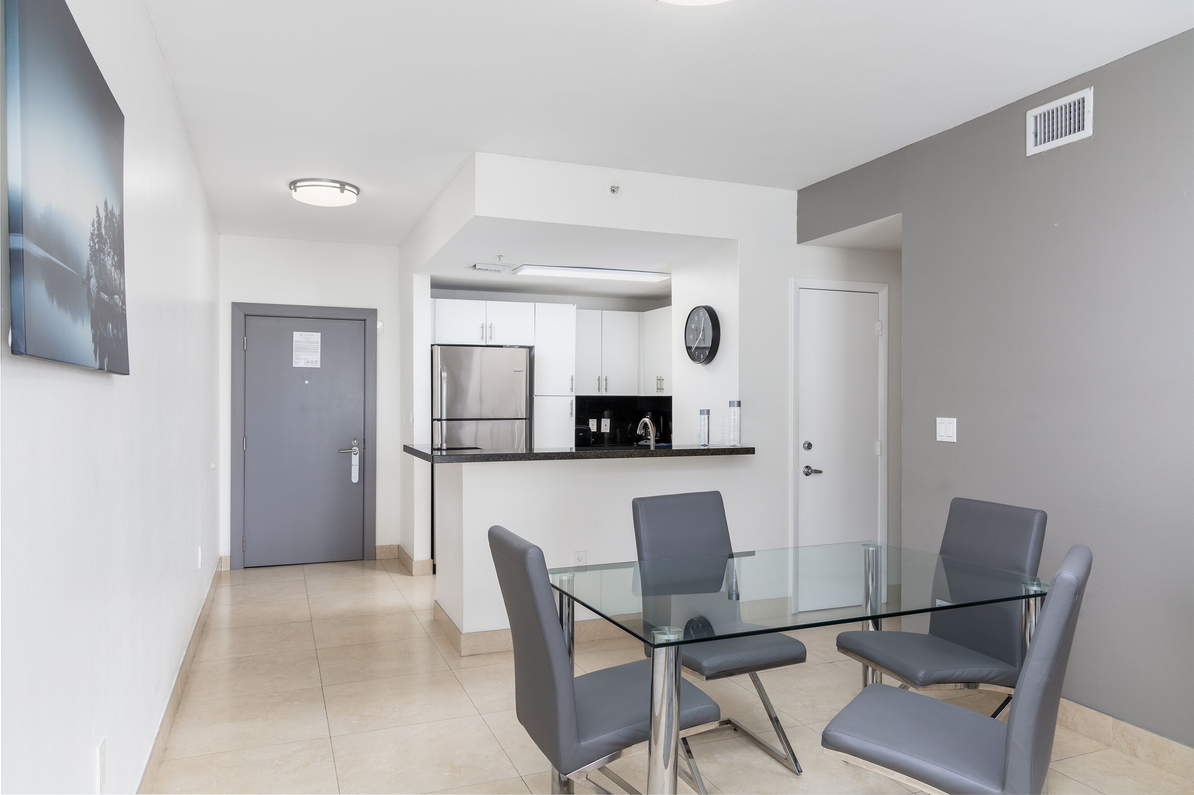 A modern kitchen with stainless steel appliances, a glass dining table with four chairs, and a grey wall.
