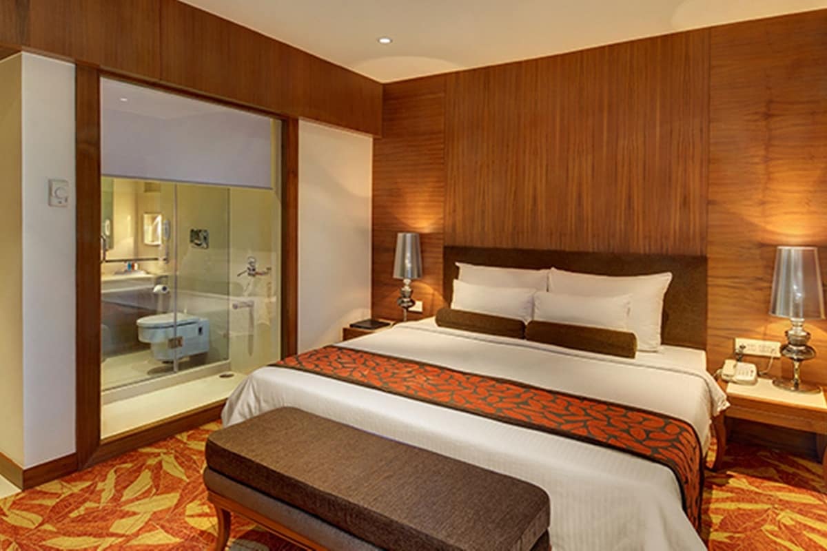 A hotel room with a bed, bedside lamps, a nightstand, a bench, a bathroom with a glass shower enclosure, and wooden paneling on the walls.