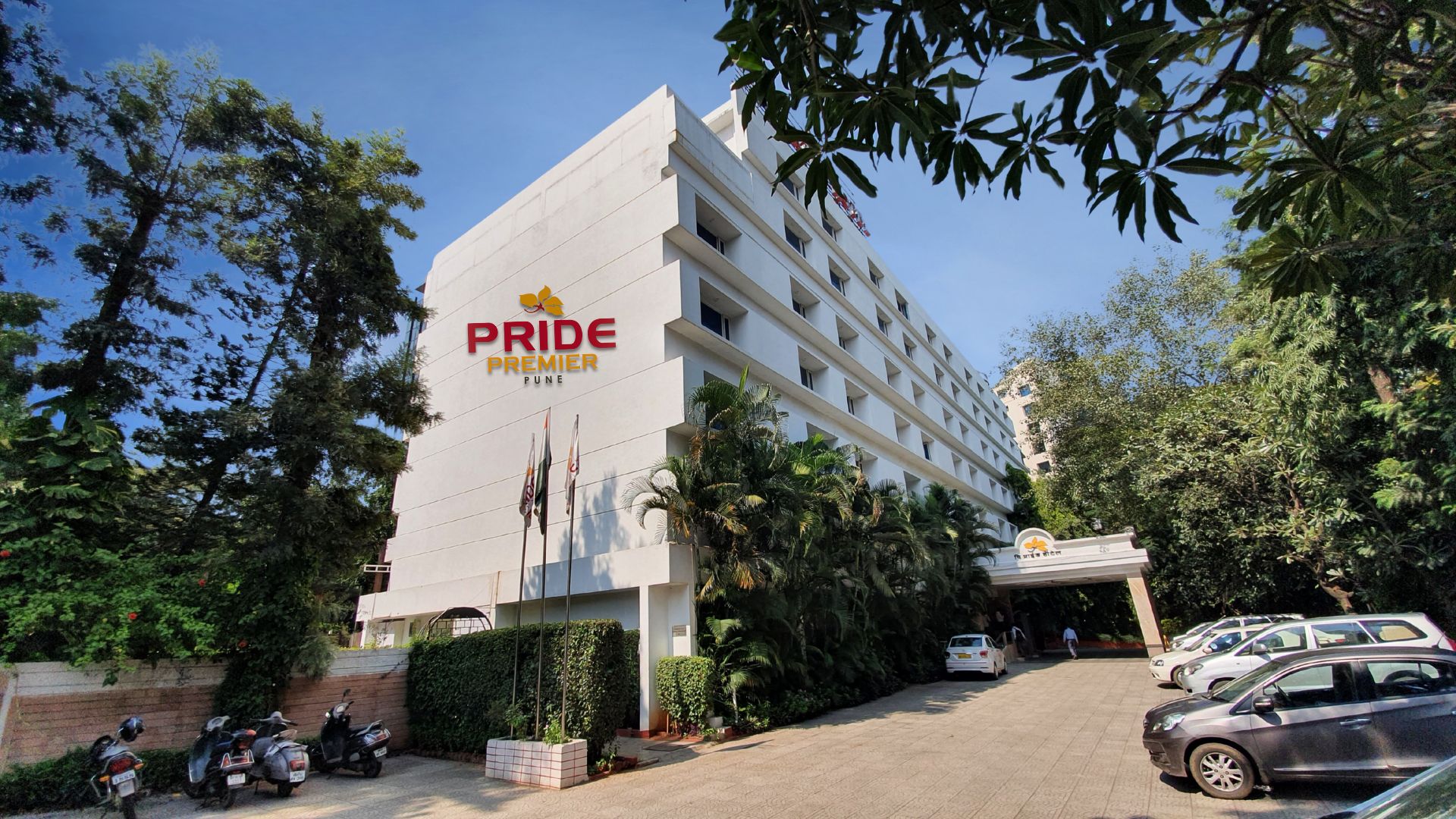 A multi-story building with the signage 'PRIDE PREMIER PUNE' indicating a hotel. There are parked cars and a few motorcycles in front.