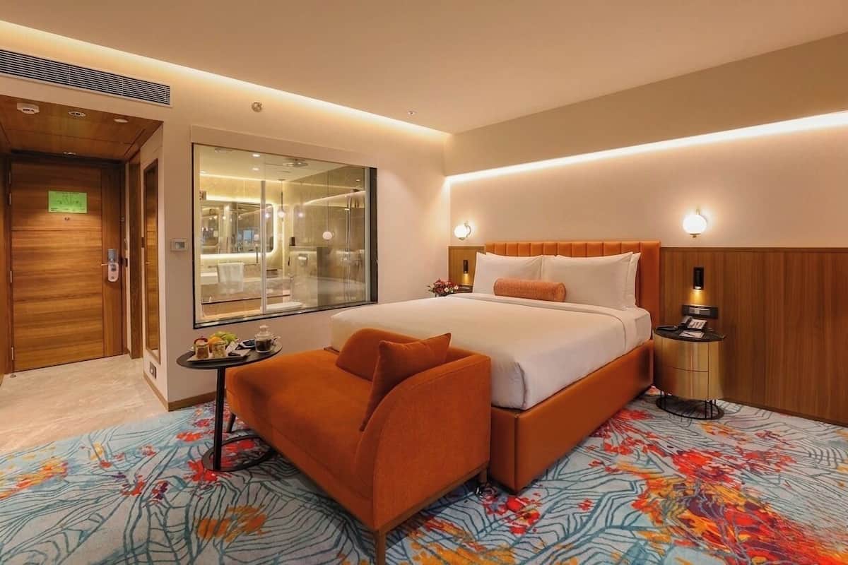 A hotel room with a bed, a sofa, a small table with amenities, a mirror, and a bathroom visible through a glass door.