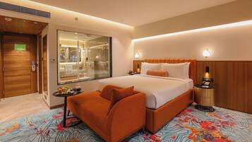 A hotel room with a bed, a sofa, a small table with amenities, a mirror, and a bathroom visible through a glass door.