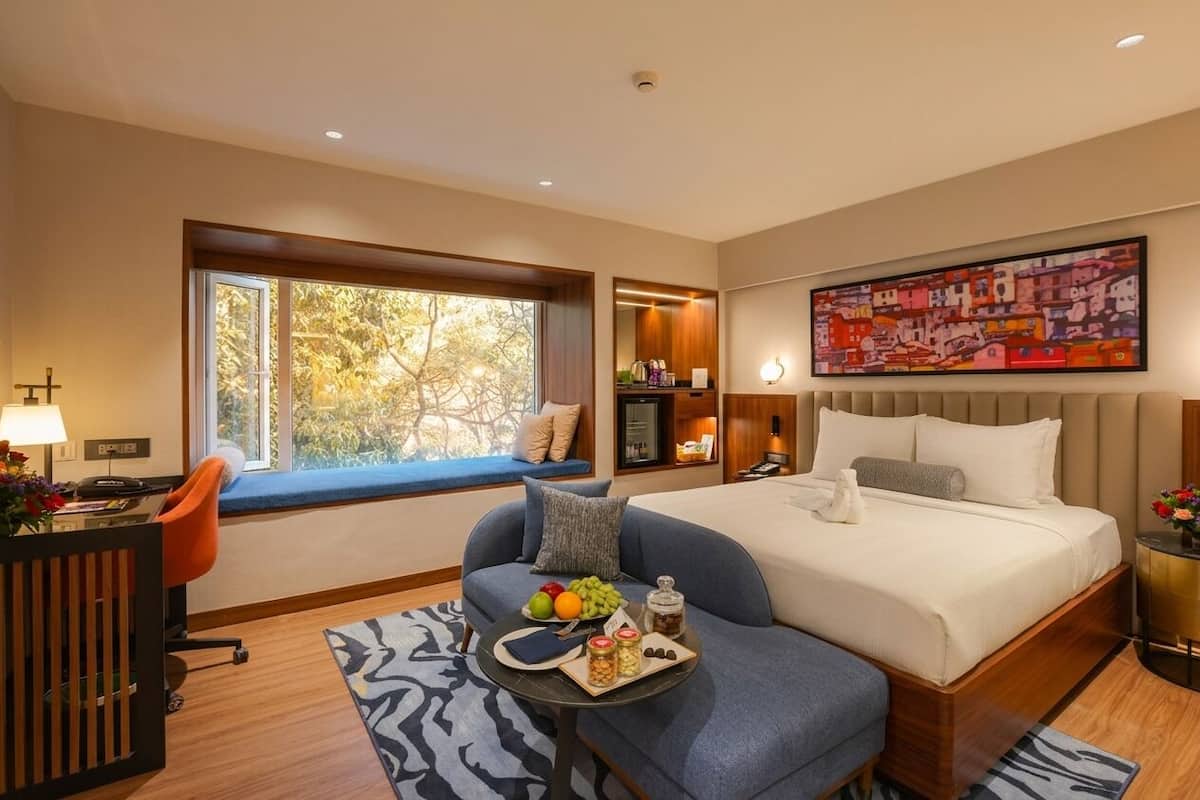A modern hotel room with a large bed, a desk, a chair, a small table with snacks, a window with a view, and a painting on the wall.