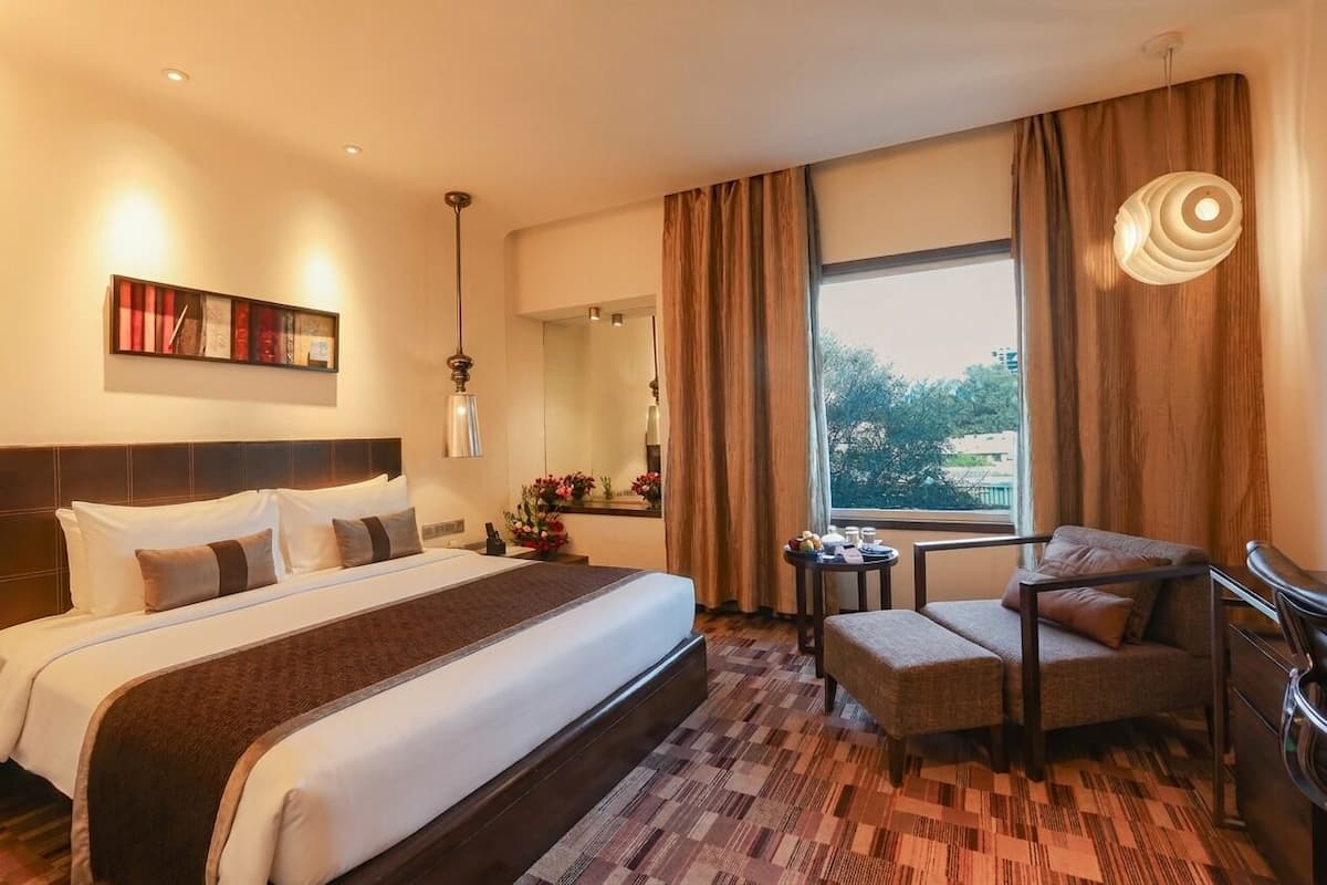 A hotel room with a large bed, a chair, a small table, a painting, and a window with curtains.