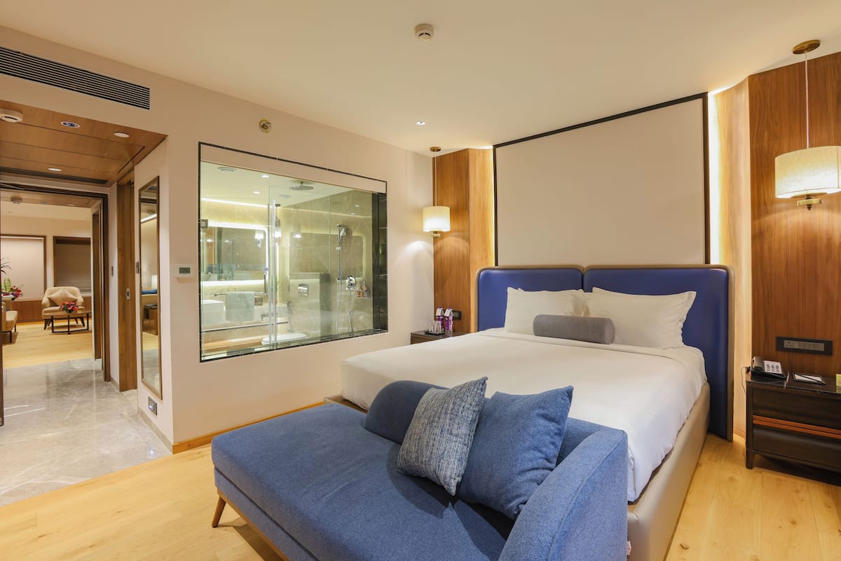 A modern hotel room with a large bed, a sofa, a glass shower area, and a wooden panel wall.