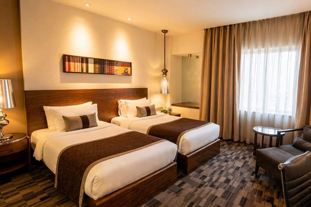 A hotel room with two beds, a desk, a chair, a lamp, a mirror, and a window with curtains.