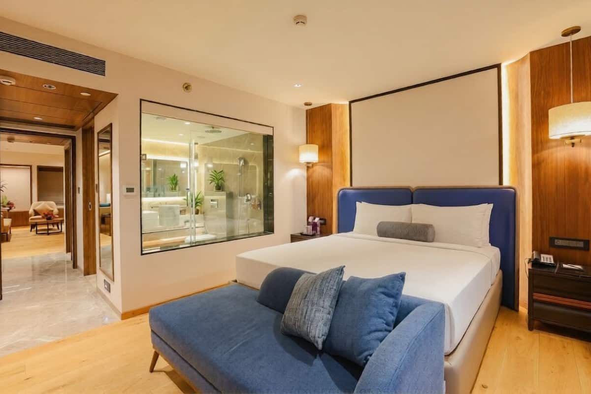 A modern hotel room with a large bed, a sofa, a glass wall, and a wooden accent wall.