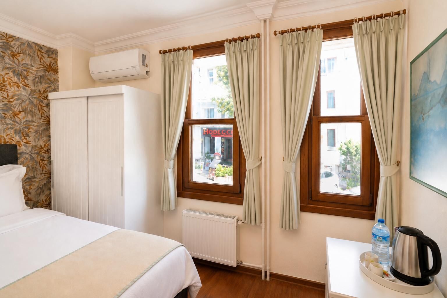 Habitación de hotel con cama, radiador, ventana con cortinas, radiador en la pared y mesa con hervidor y botella de agua.