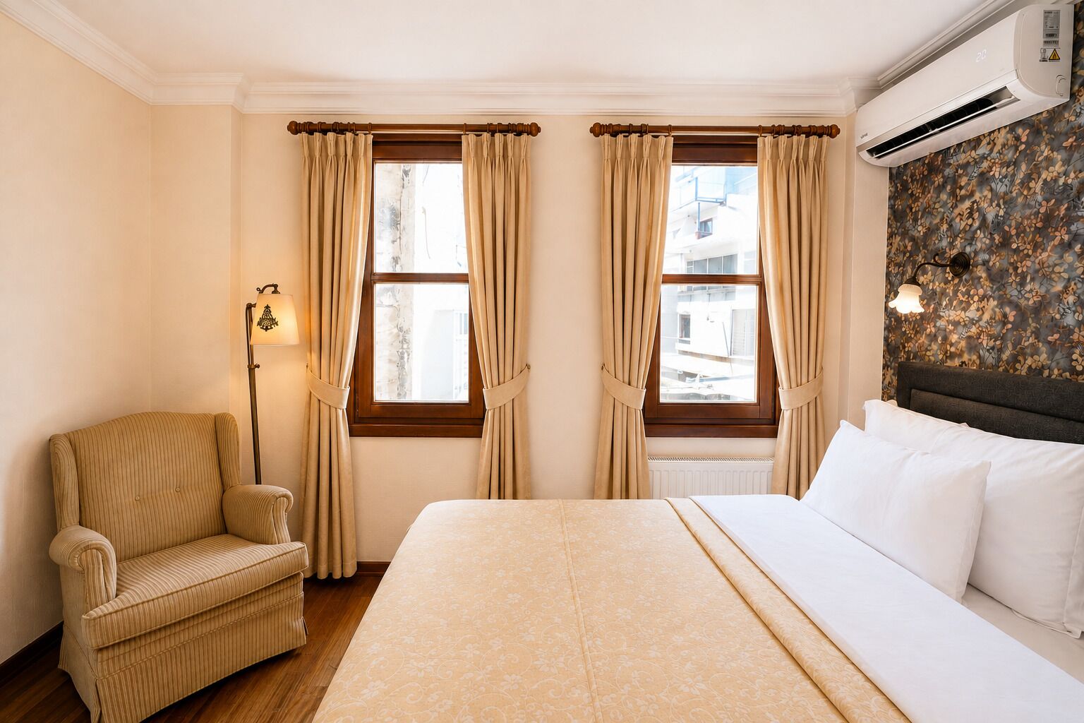 Habitación de hotel con cama, una silla, dos ventanas con cortinas, un equipo de aire acondicionado y una lámpara de pared.