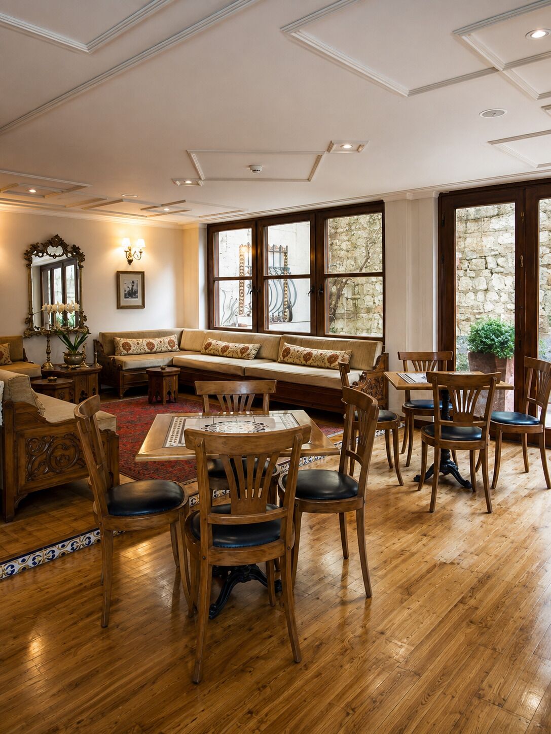 A room with a wooden floor, a sofa, a dining table with chairs, and large windows with stone walls.