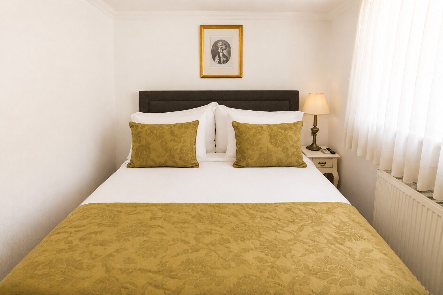 Ett hotellrum med en säng, två kuddar, ett sängbord med en lampa och en inramad tavla på väggen.