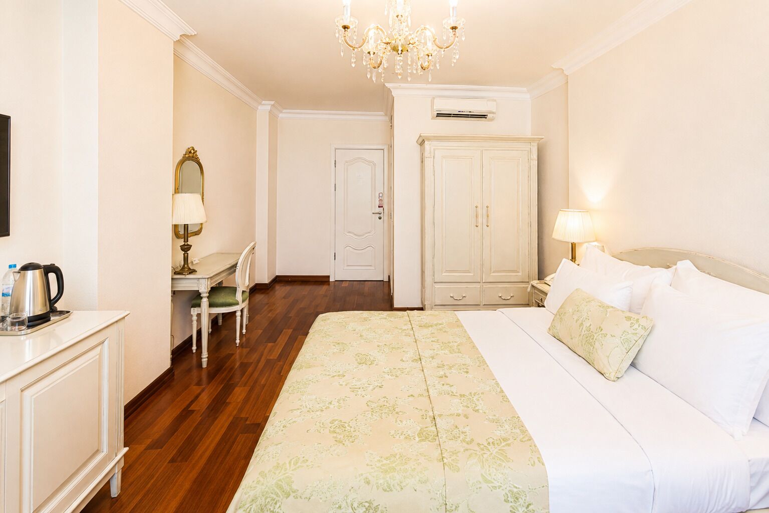 A hotel room with a bed, a desk with a chair, a wardrobe, and a chandelier.