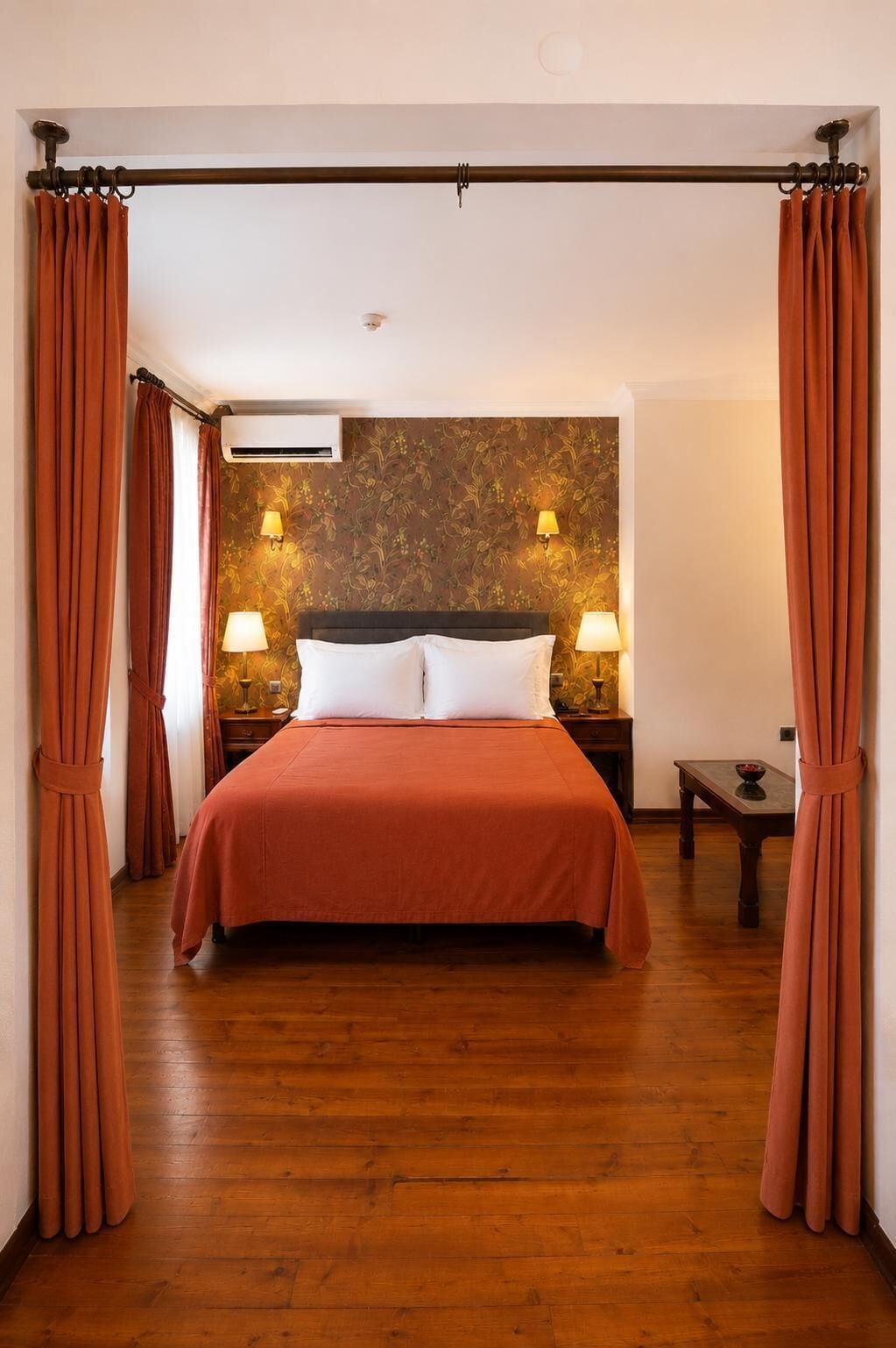 A bedroom with a bed, curtains, two bedside lamps, a nightstand, and a wall-mounted air conditioner.