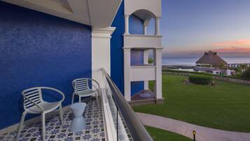 Un balcón con dos sillas blancas y una mesa pequeña, con vistas a un césped cuidado y una estructura con techo de paja.