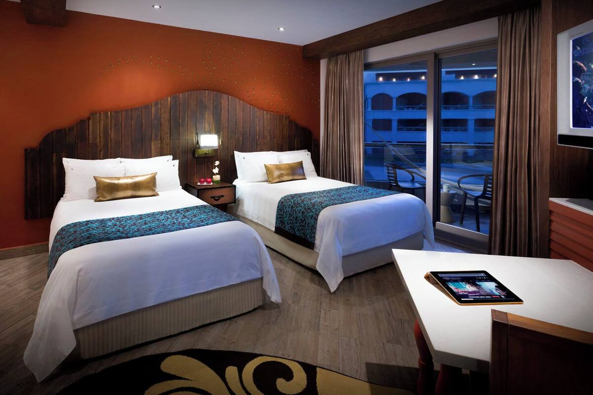 A hotel room with two beds, a desk, a tablet, and a balcony with a view of the city.