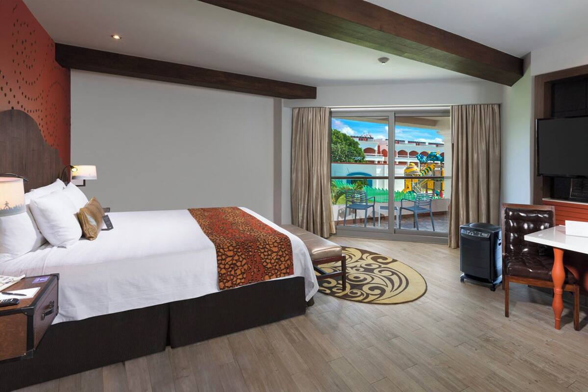 A hotel room with a bed, a desk, a chair, a TV, and a balcony with a view of a playground.