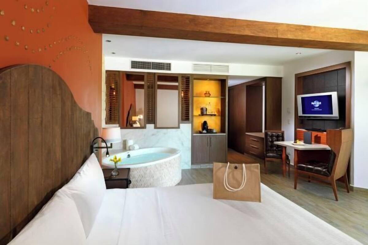 A modern hotel room with a large bed, a sitting area with a TV, a bathroom with a bathtub, and a kitchenette.