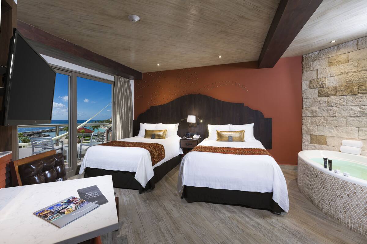 A hotel room with two beds, a view of the ocean, a stone wall, and a bathtub.