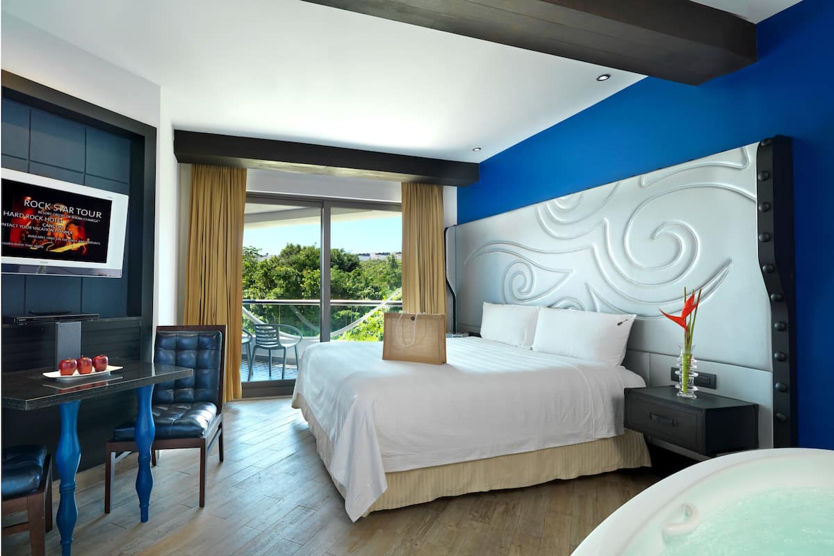 A modern hotel room with a large bed, a TV, a dining table, and a balcony with a view.