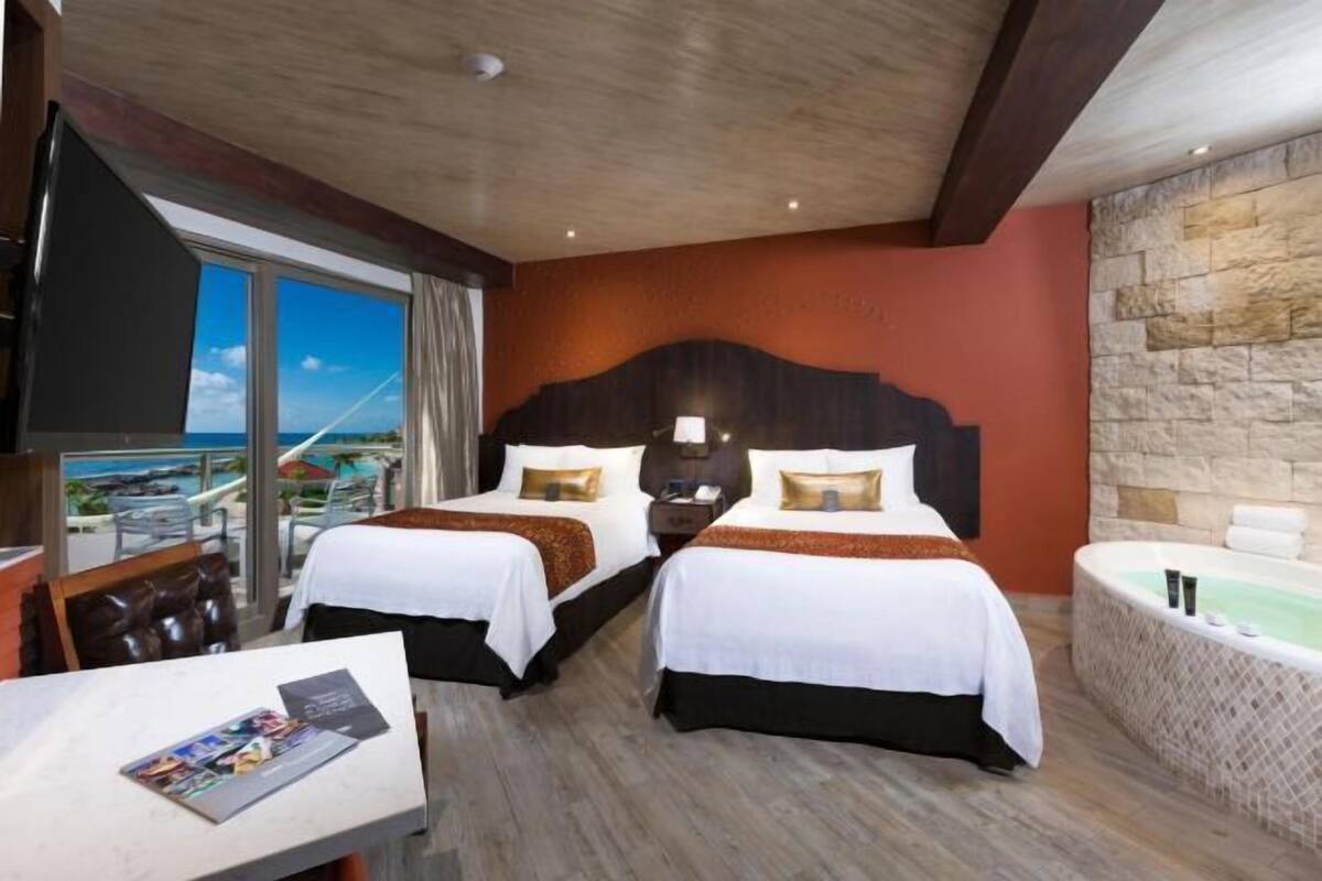 A hotel room with two beds, a large window with a view, a bathtub, and a table with a magazine.