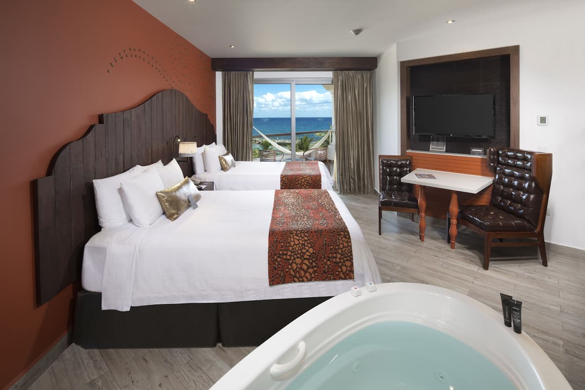 A hotel room with a large bed, a separate sofa bed, a desk with chairs, a TV, and a view of the ocean.