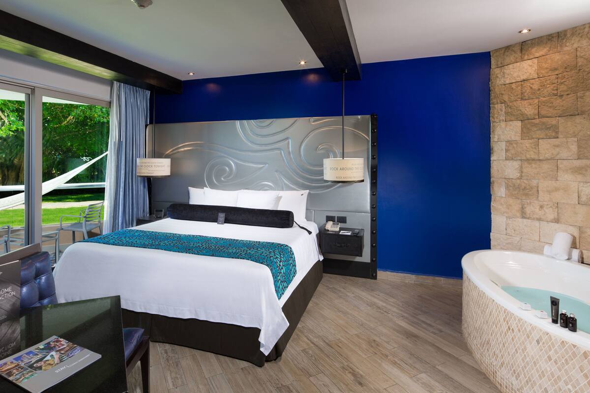 A modern hotel room with a large bed, a sitting area with a glass table, a bathtub, and a view of greenery outside.