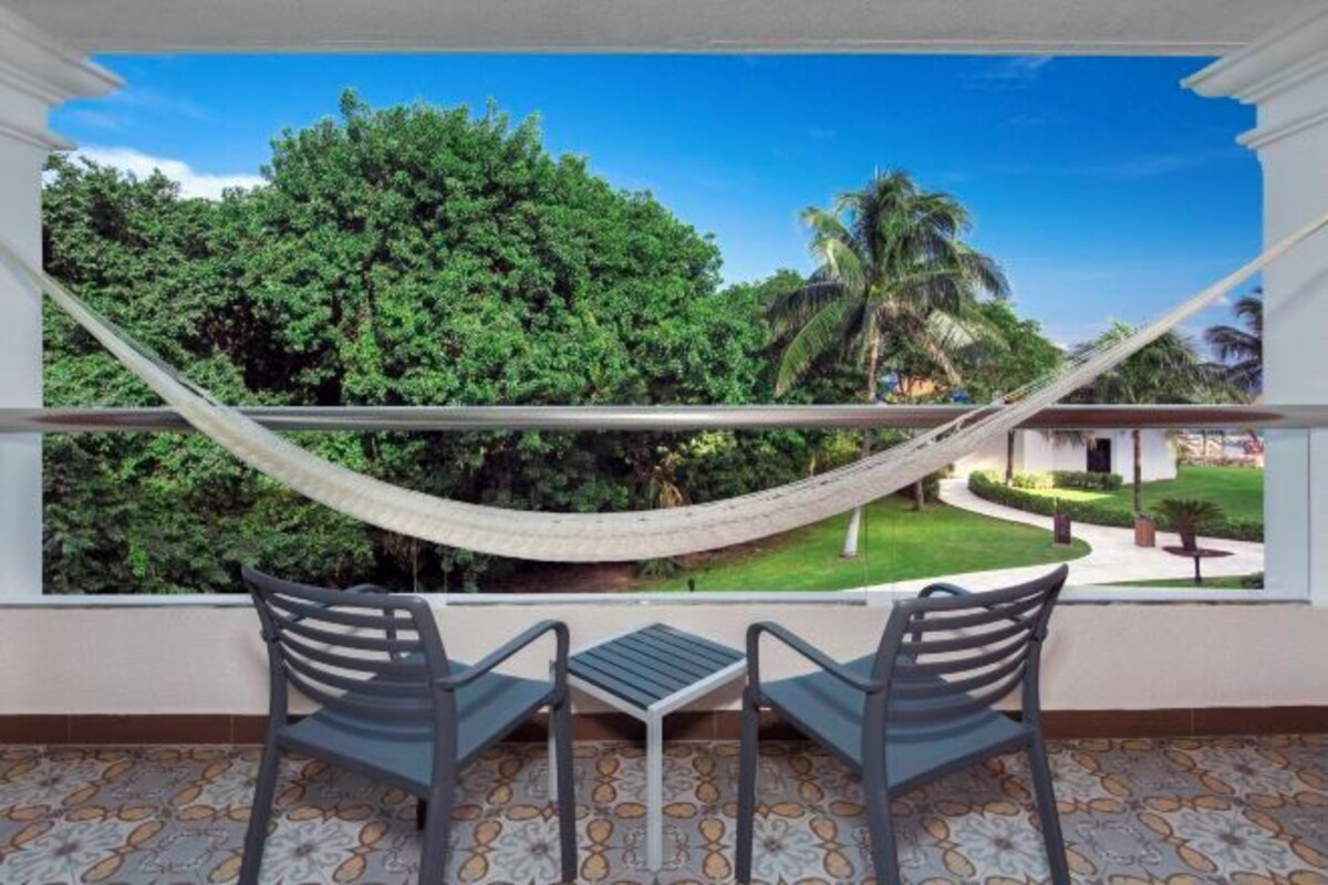 A balcony with a view of a lush garden, a hammock, and two chairs.