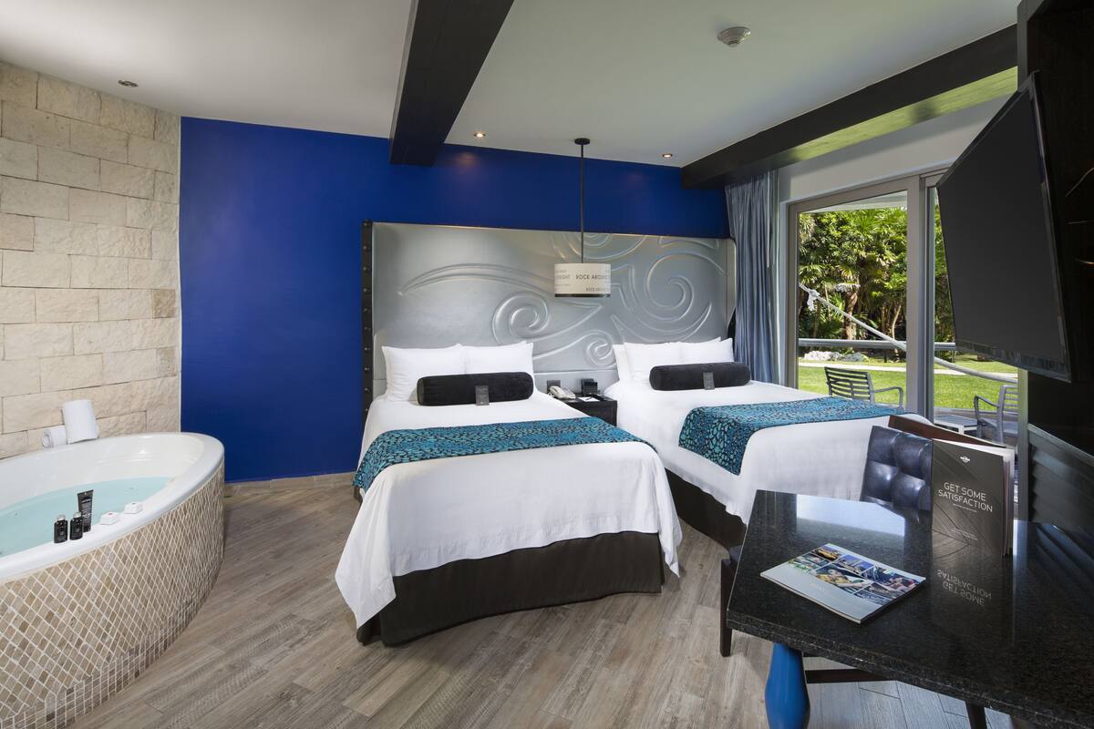 A hotel room with two beds, a large window, a bathtub, and a table with a magazine.