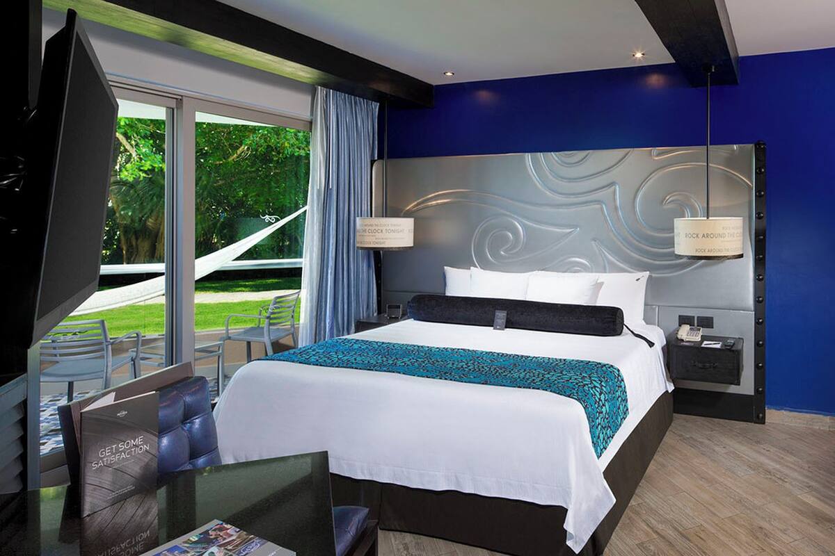 A modern hotel room with a large bed, a desk, and a TV. There is a view of greenery outside the window.