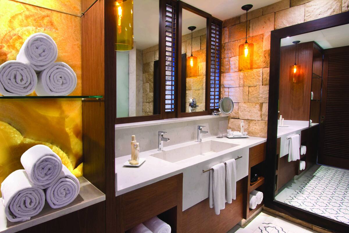 A modern bathroom with a large mirror, two sinks, and shelves holding rolled towels.