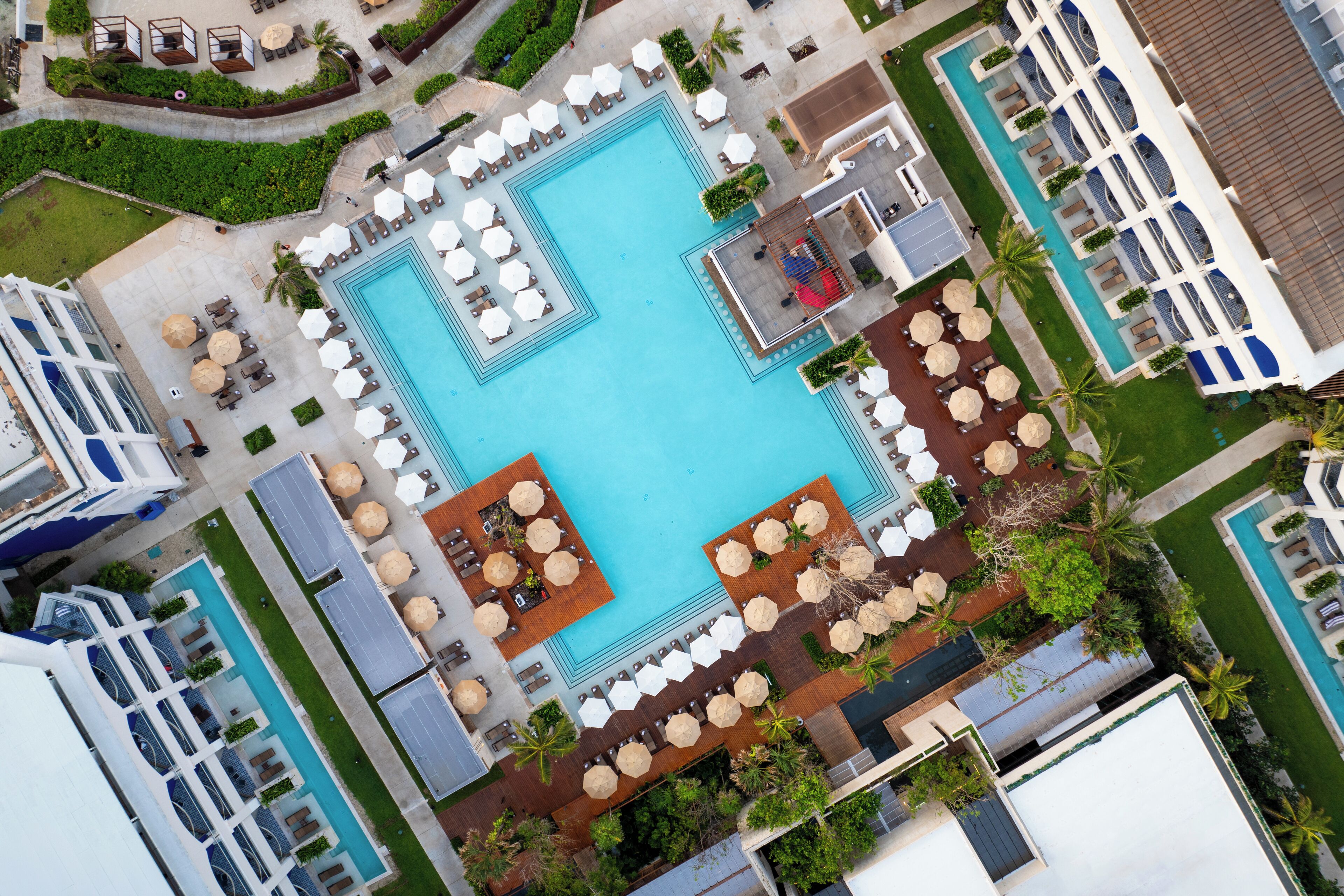 An aerial view of a hotel with a central swimming pool surrounded by sun loungers and umbrellas, multiple poolside dining areas, and lush landscaping.