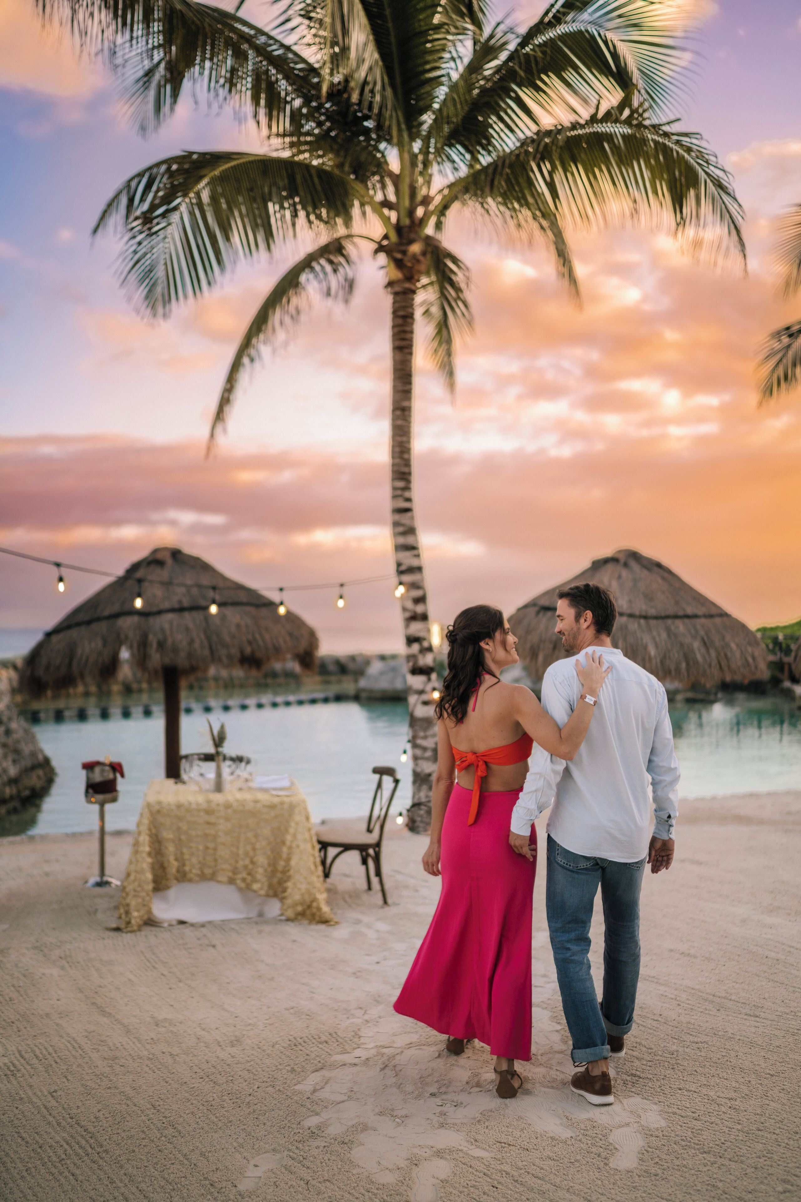 A couple walking on a beach with palm trees and a thatched-roof structure in the background.