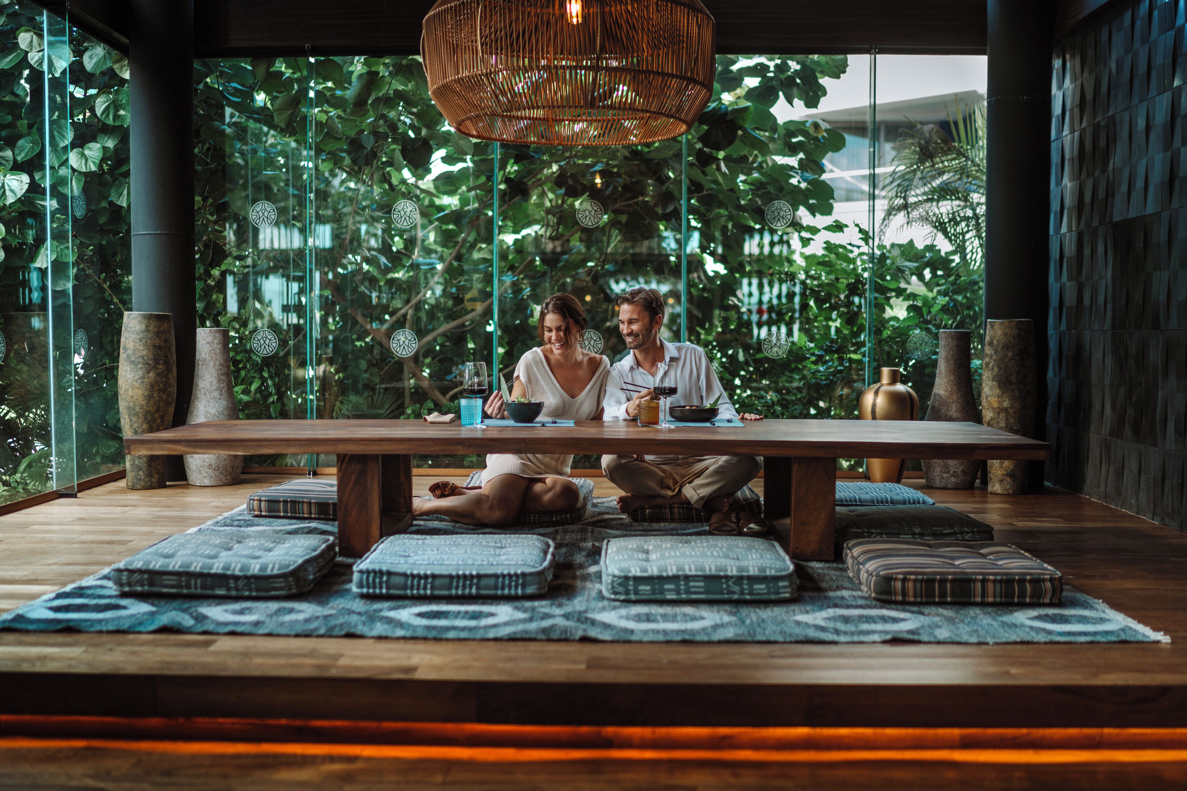 Una pareja cenando en una mesa de madera con cojines, rodeada de grandes ventanales y follaje.