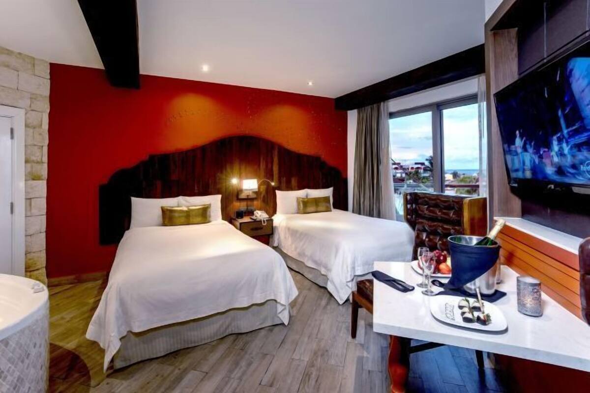 A hotel room with two beds, a TV, a table with a bottle of champagne, and a view of the city.