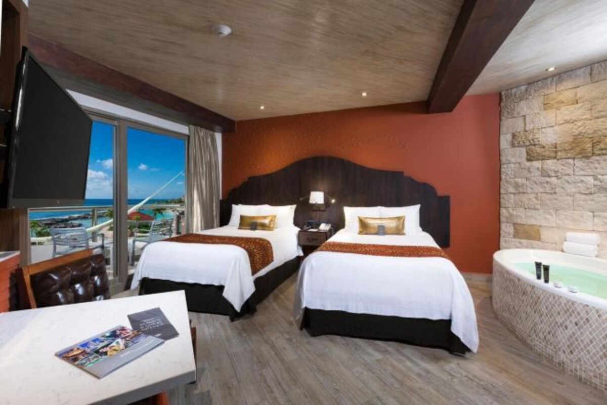 A hotel room with two beds, a large window with a view, a bathtub, and a table with a magazine.