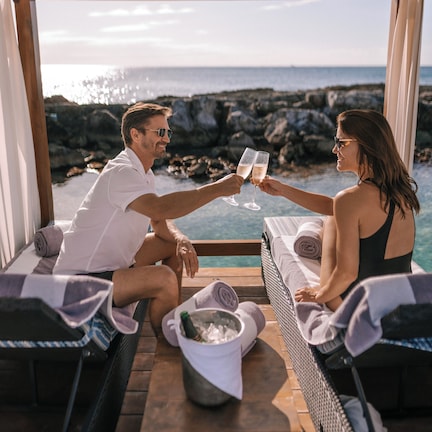 Una pareja brindando con copas de champán en una zona de estar exterior acogedora con vistas al océano.