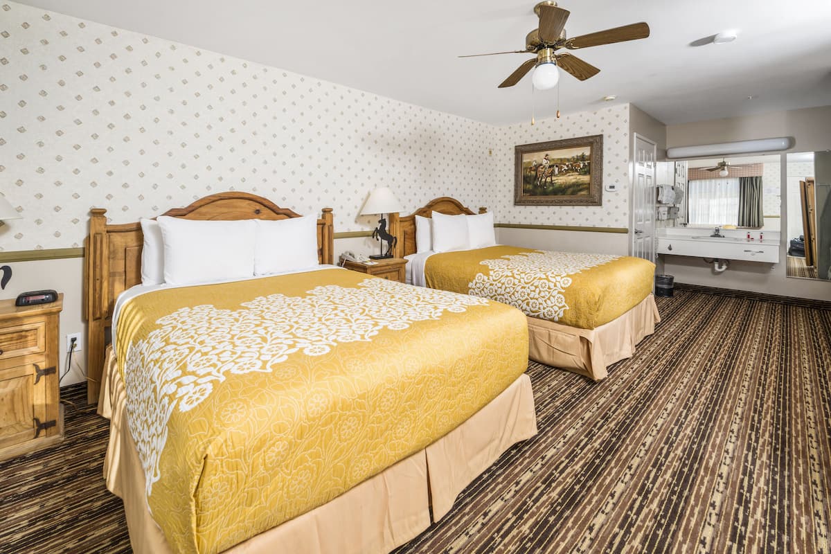 A hotel room with two beds, a ceiling fan, a painting on the wall, and a bathroom with a mirror and sink.