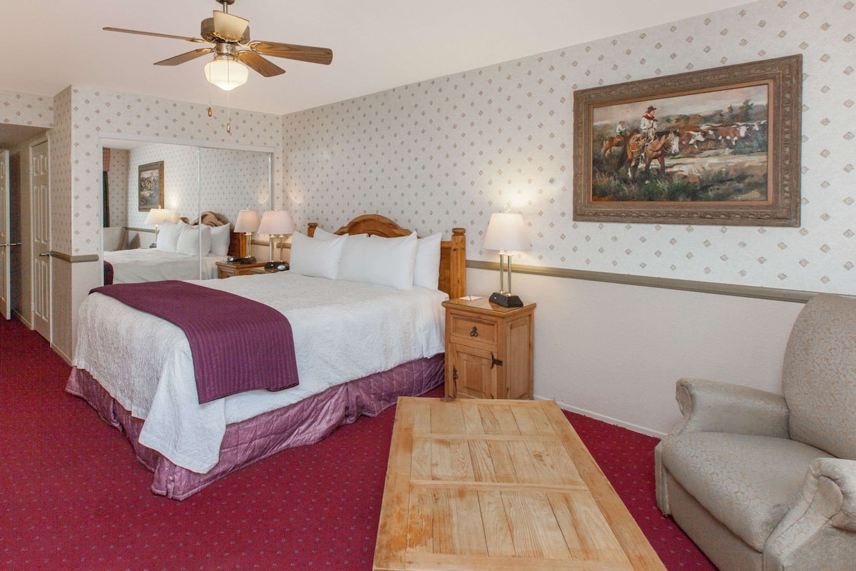 A hotel room with a large bed, a wooden nightstand, a chair, a ceiling fan, and a painting on the wall.