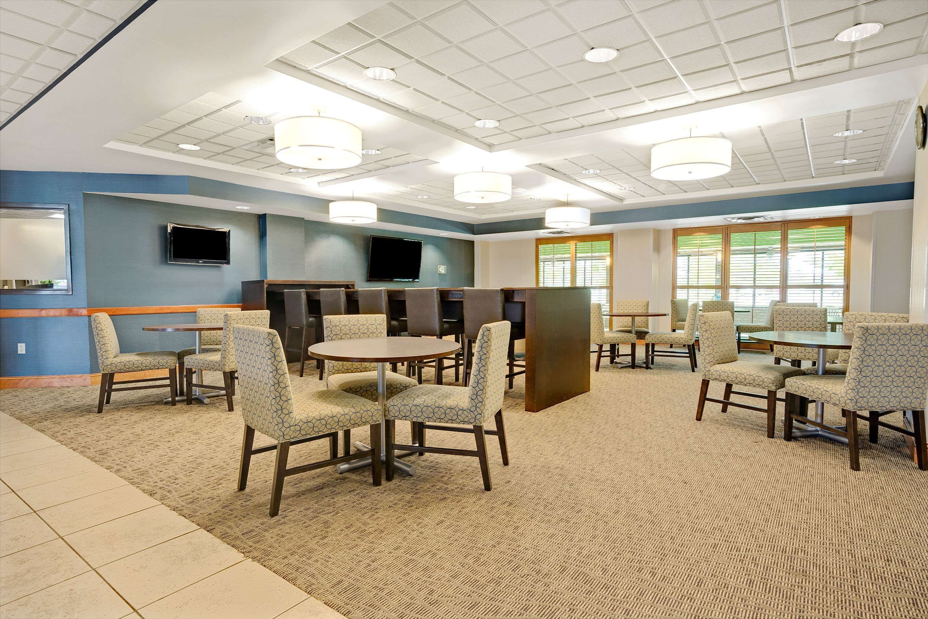 A spacious indoor area with multiple seating arrangements, including round tables and chairs, a flat-screen TV mounted on the wall, and large windows allowing natural light.