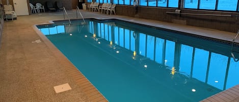 An indoor swimming pool with clear water, surrounded by a tiled deck and a brick wall. There are chairs and a bench near the pool, and a window with a view of a snowy landscape.
