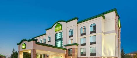 A multi-story hotel with a prominent green and white sign, a covered entrance, and a parking area.