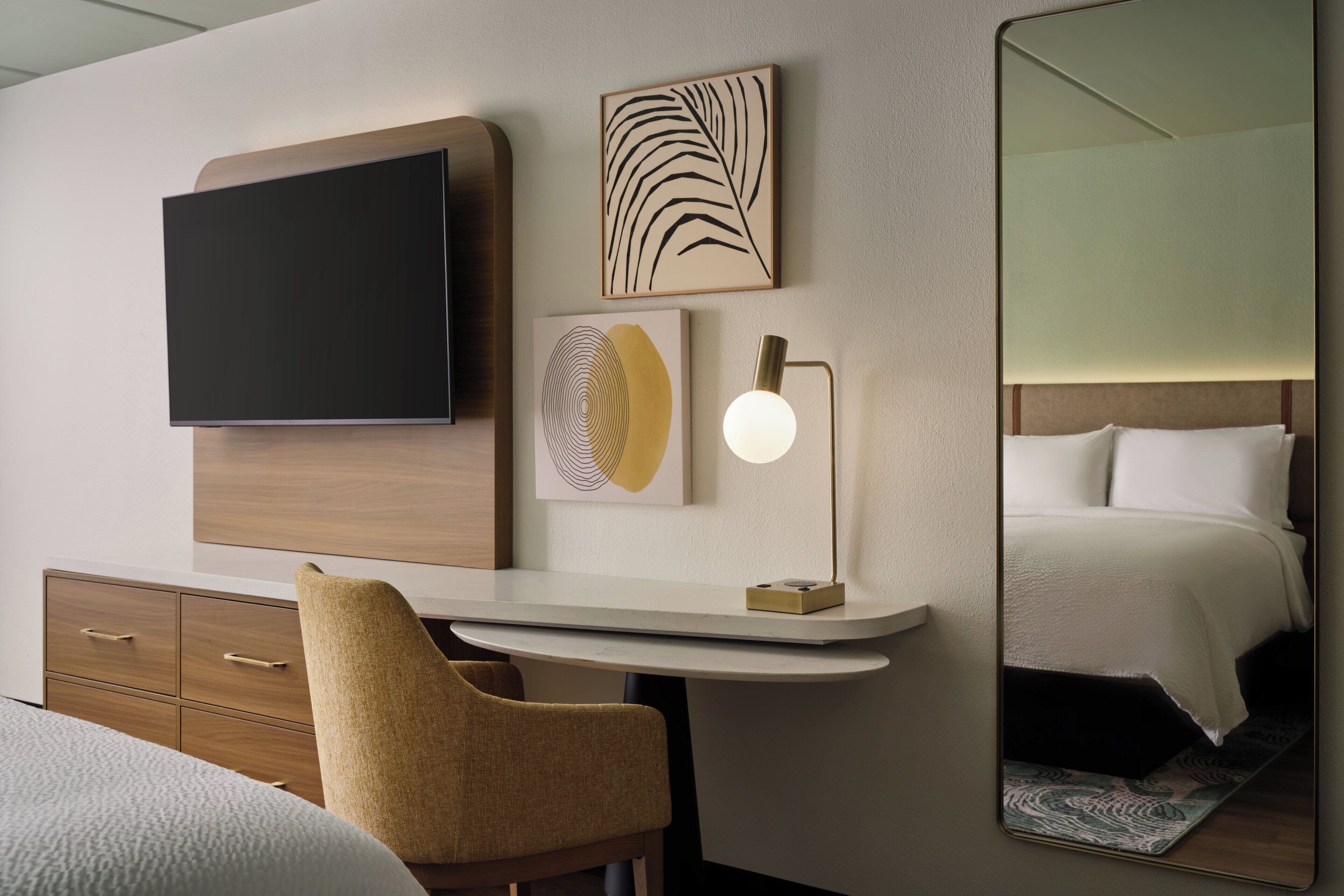A hotel room with a flat-screen TV, artwork, a desk with a lamp, a chair, and a bed with a headboard.