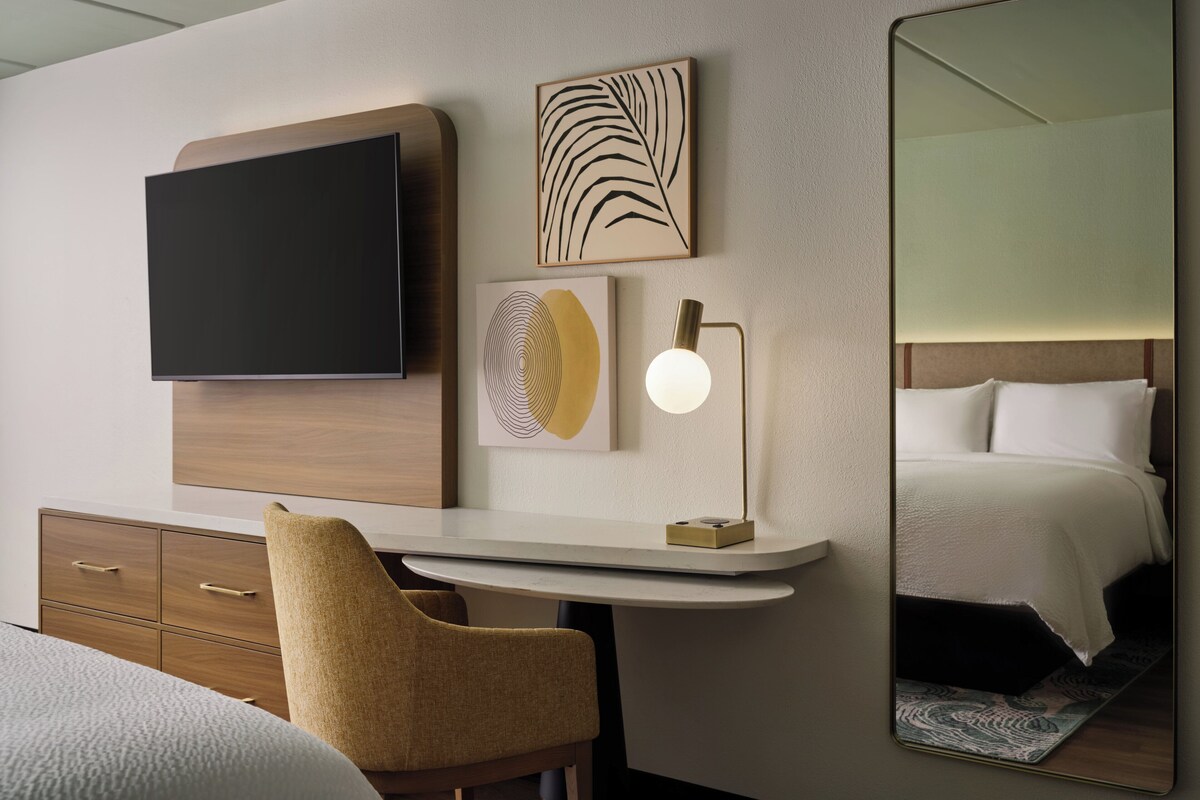 A hotel room with a flat-screen TV, artwork, a desk with a lamp, a chair, and a bed with a headboard.