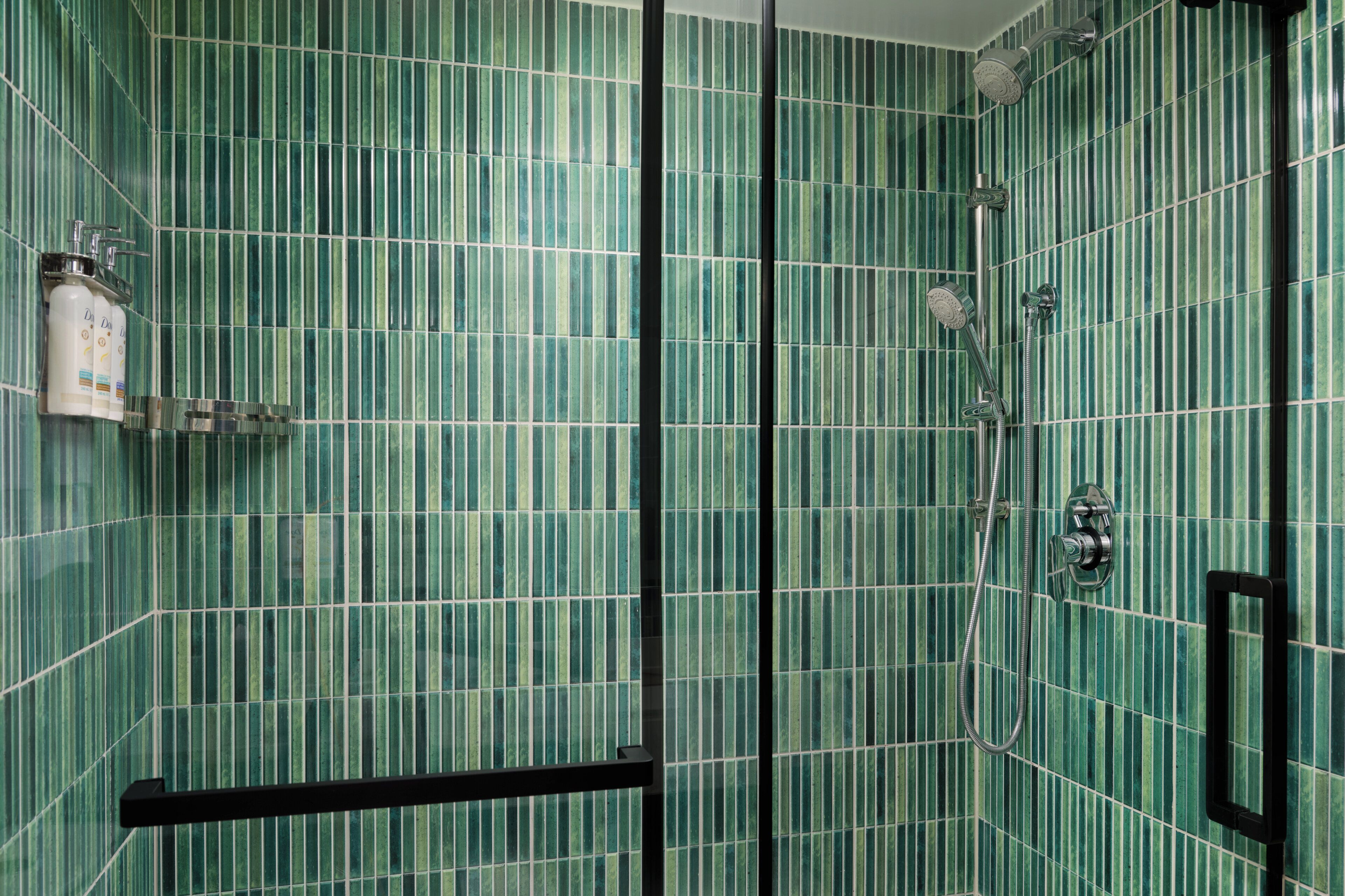 Un baño con baldosas verdes a rayas, ducha con cabezal de ducha manual y dispensador de jabón en la pared.