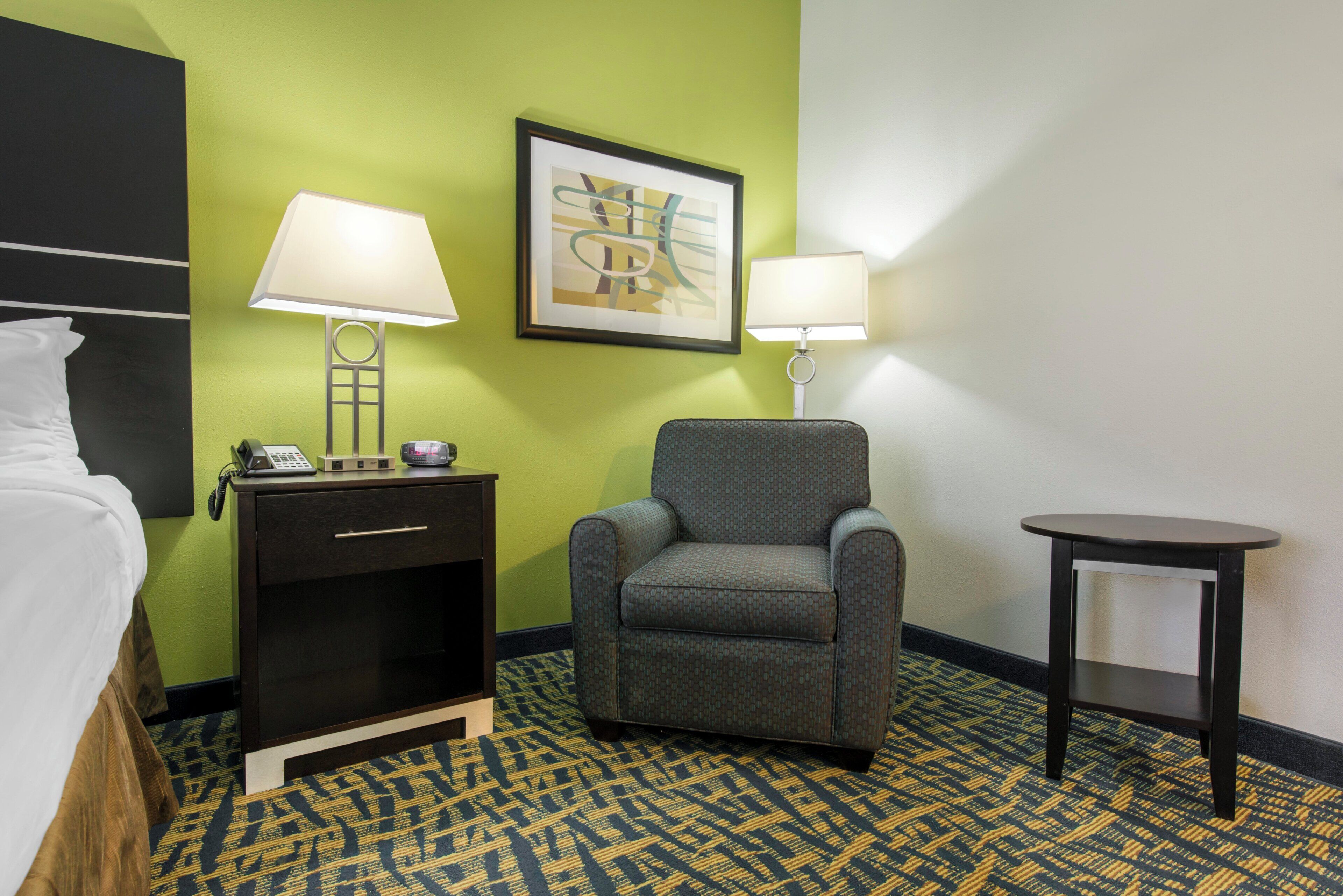 A hotel room with a bed, a chair, a desk, a lamp, and a framed picture on the wall.