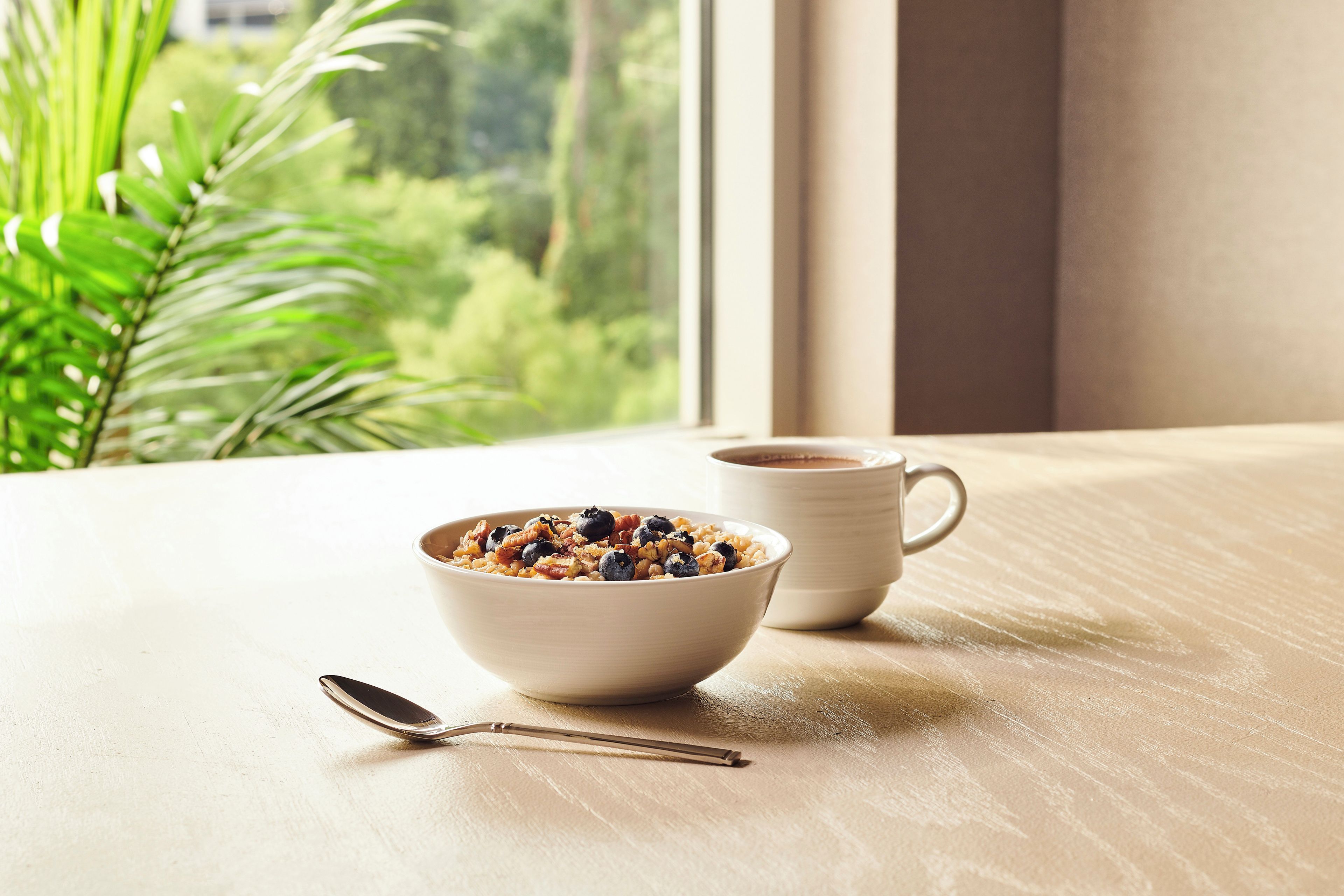 A bowl of cereal with a spoon on a wooden table by a window with a view of greenery.